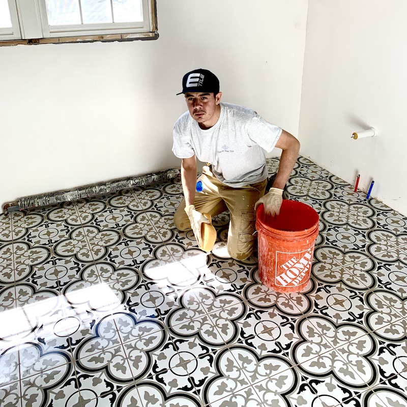 Contractor kneeling on a freshly installed patterned tile floor, holding a grout sponge and an orange bucket, showcasing precision and attention to detail during tile installation.